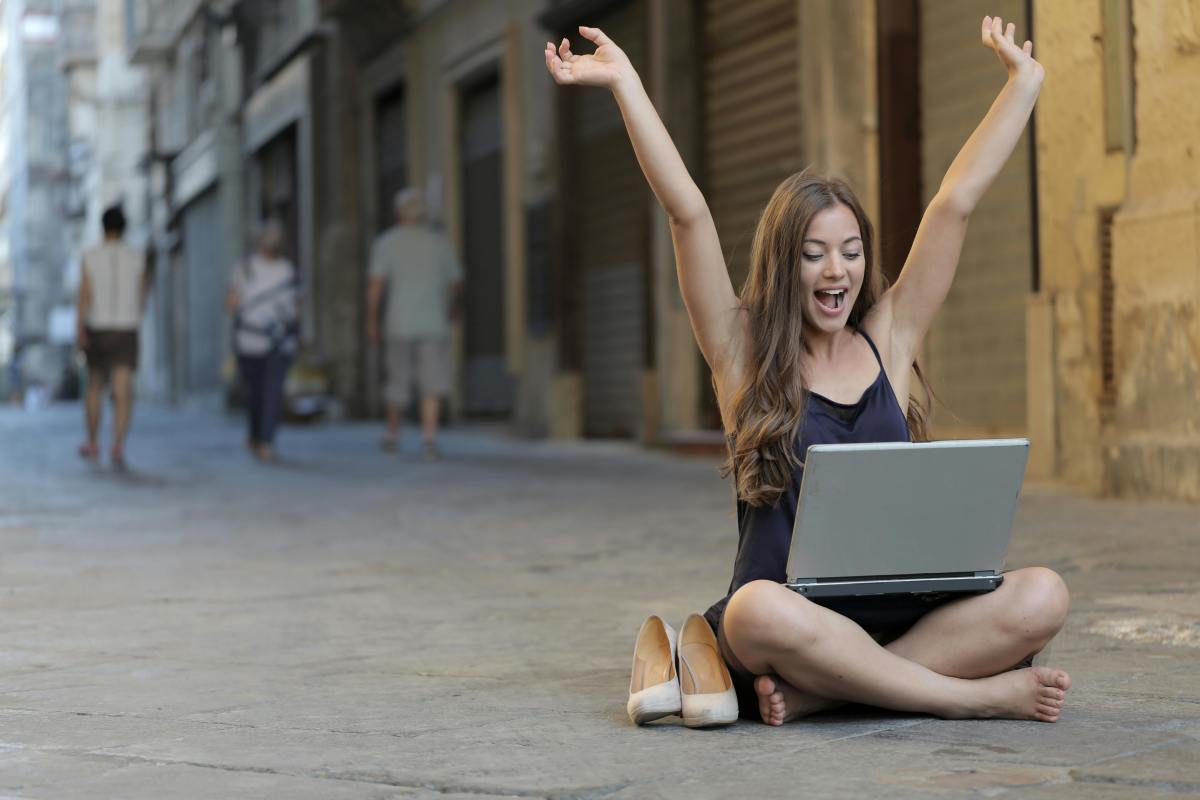 Woman raising her hands up while sitting on the street with a laptop on her lap